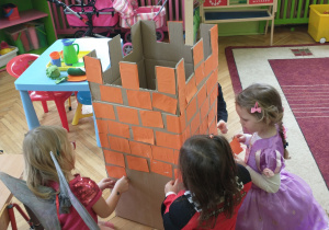 Trzy dziewczynki w bajkowych strojach przyklejają cegiełki do zamku